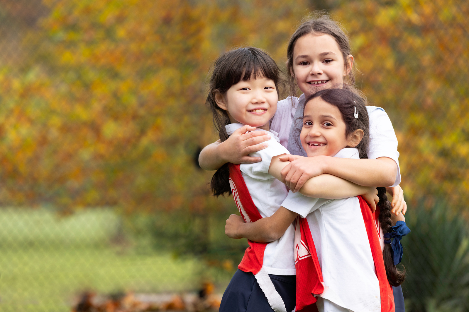 Three young girls wearing netball bibs hug each other in front of autumnal leaves on a court at a private school in the Dorset countryside.