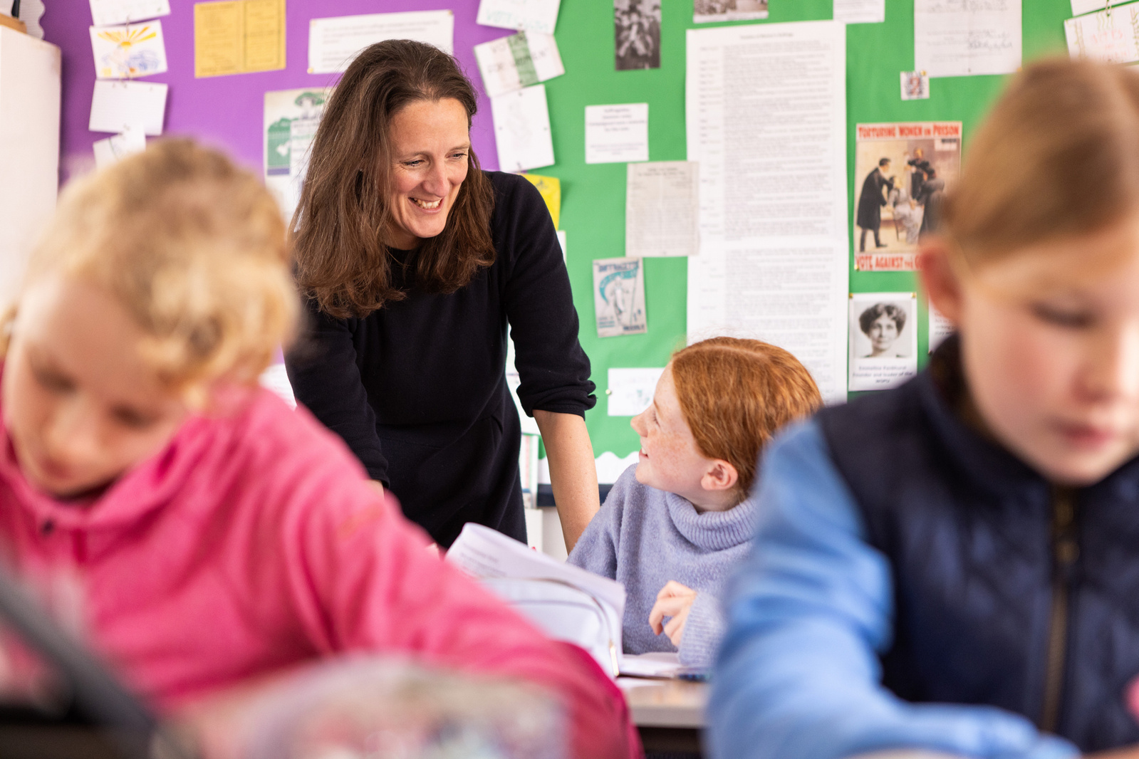 A teacher and a pupil look at each other in an independent school classroom in South England. The teacher is standing over the student and two more students are blurred in the foreground writing.