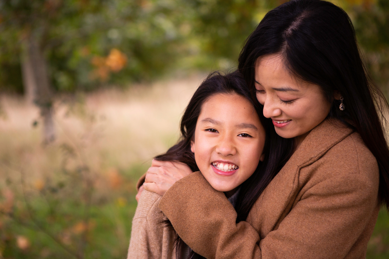 A mother and daughter hug. The daughter is looking straight at the camera and the mother is looking at her daughter. They are in Moors Valley Country Park.