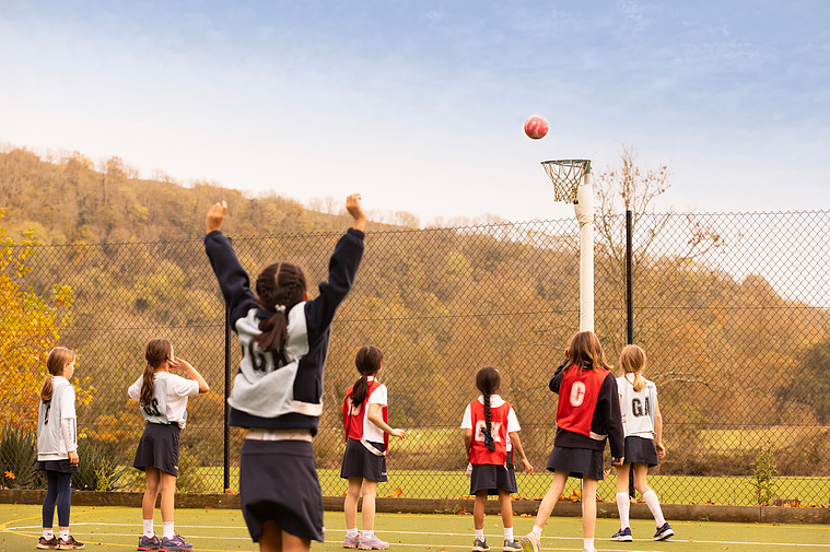A girl celebrates scoring a goal at a netball match in the countryside at a private school. The ball is about to go in the net and 7 children are watching it go in.