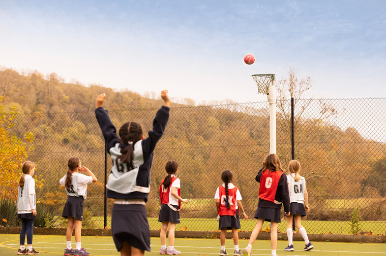 A girl celebrates scoring a goal at a netball match in the countryside at a private school. The ball is about to go in the net and 7 children are watching it go in.