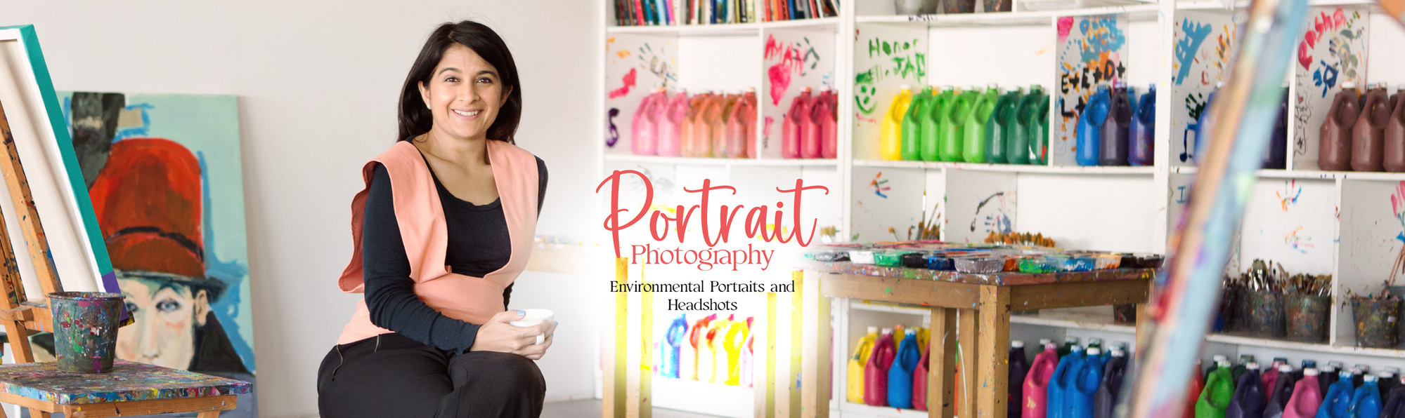 A creative business owner sits in her studio in Dorset surrounded by colourful paint and easels. She is looking at the camera and holding a mug. 