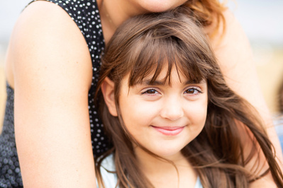 A daughter with long brown hair, in her mother's arms, smiles at the camera at a beach on the Jurassic Coast.
