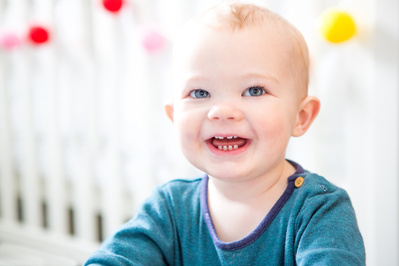 A baby sits in front of his cot in Bath, UK with a big smile. Colourful pompoms decorate the cot behind him.