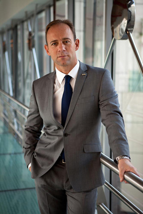 A business man poses for a portrait in a grey suit and black tie with a white shirt. He is in a corridor in an office building in Poole, Dorset.