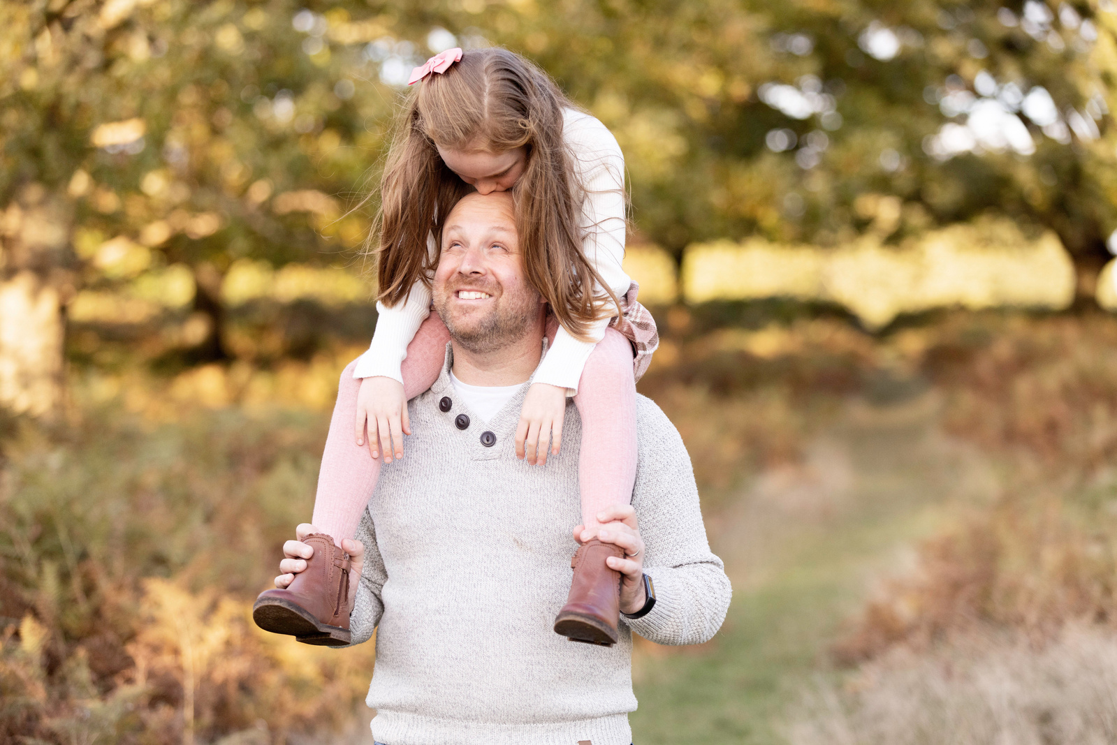 A Father carries his 7 year old daughter on his shoulders. She is looking down at him and he's looking up at her. They are in the countryside.