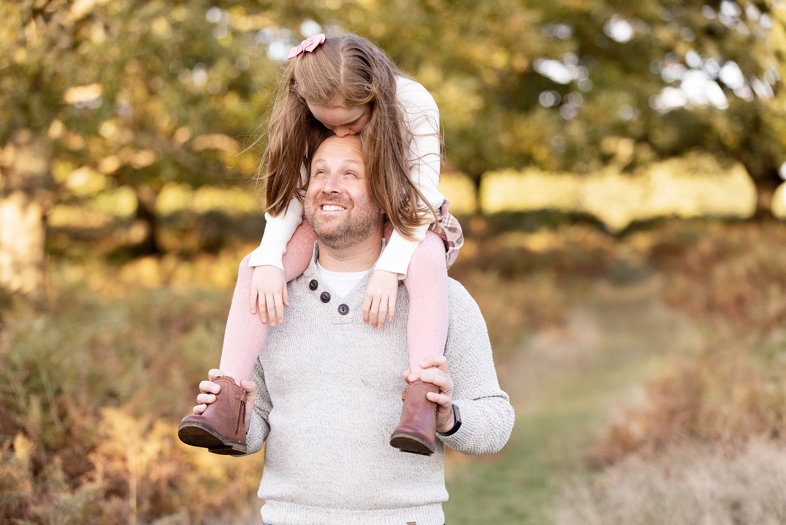 A Father carries his 7 year old daughter on his shoulders. She is looking down at him and he's looking up at her. They are in the countryside.