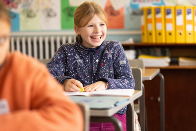 A girl sits at a desk in a classroom and smiles. She is sitting behind another pupil and has a row of yellow folders behind her. 
