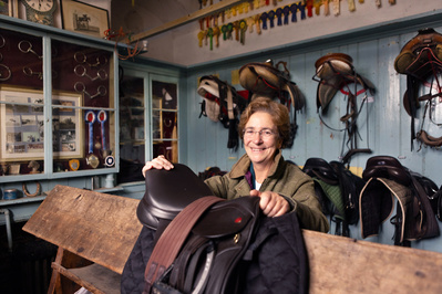 The Headmistress of Hanford Girls school poses in the tack room with a saddle. She is surrounded by saddles and rosettes.