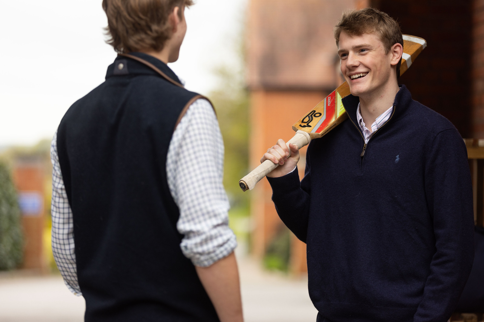 Two boys at Clayesmore School Dorset chat. One is holding a cricket bat over his shoulder.