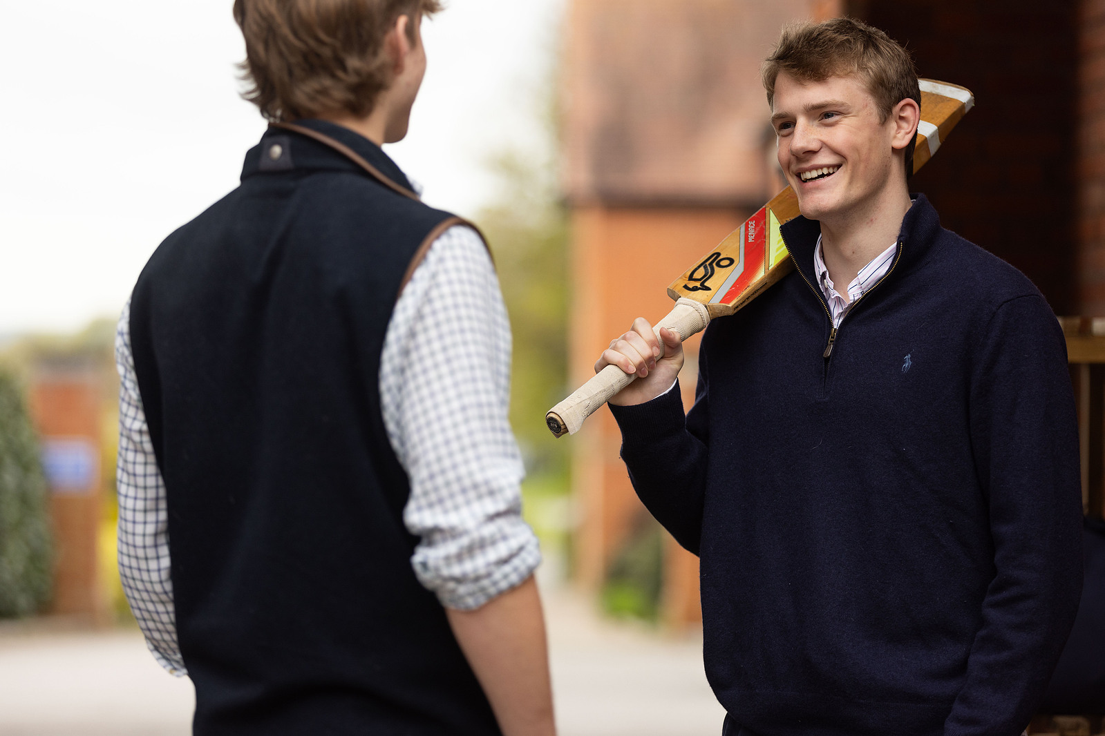Two boys at Clayesmore School Dorset chat. One is holding a cricket bat over his shoulder.