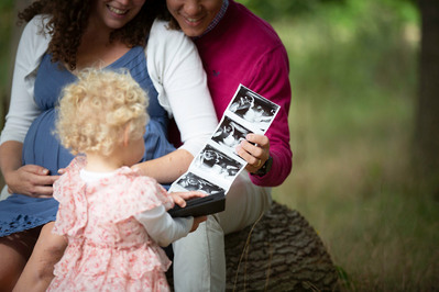 A pregnant lady and her partner sit on a log in a forest and show their daughter x-ray images from her pregnancy scan.