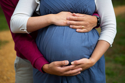 A close up shot of a pregnant lady, just showing her tummy. Her arms are clasped under her tummy and her partners arms are above it. He is standing behind her but you can't see either of their faces. At the beach on the Jurassic Coast.