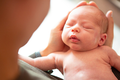 A Father holds his newborn baby's head in his hands. The baby's head fills his whole hands. The baby is topless and asleep. They're at home in the their living room in Dorchester, Dorset.