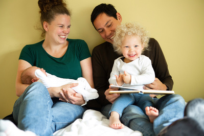 A family of four, 2 parents, a 2 year old girl and a newborn baby sit on a bed smiling. The girl is reading a book but looking straight at the camera in their bedroom at home.
