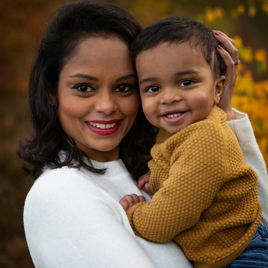 A mother in a white jumper holds her smiling 2 year old son who wears a mustard coloured jumper. They're in the New Forest.