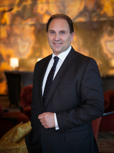 The General Manager of a hotel stands in his hotel lobby posing for an environmental portrait. He is smiling and wearing a very smart black suit, black tie and white shirt.