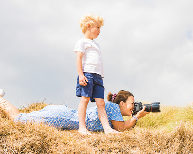 A professional photographer lies on the ground in a sand dune taking a photograph with her Canon camera. A young boy stands next to her looking at what she is shooting. At the beach in Swanage, Dorset.