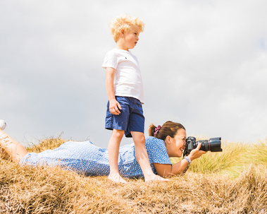A professional photographer lies in the sand dunes at Hengistbury Head and takes a photo with her Canon camera. A young boy stands next to her watching what she is photographing.