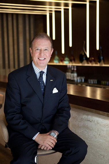 A business man sits on a bar stool in a reception in a smart navy suit and tie. He is posing for a corporate portrait and looking straight at the camera.