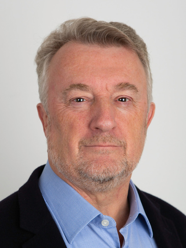 Head shot of a grey haired business man in a black jacket and blue shirt in front of a grey background.
