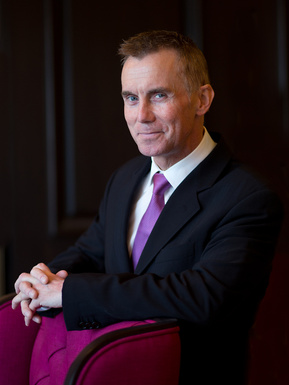 A corporate portrait of Chef Gary Rhodes. Here is posing as a business man for the camera. He's wearing a black suit, white shirt and purple tie and is smiling slightly.