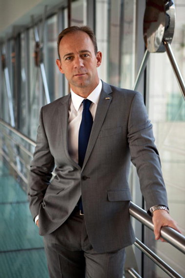 A business man in a grey suit poses for a corporate portrait. He looks very serious and has his hand in his pocket.