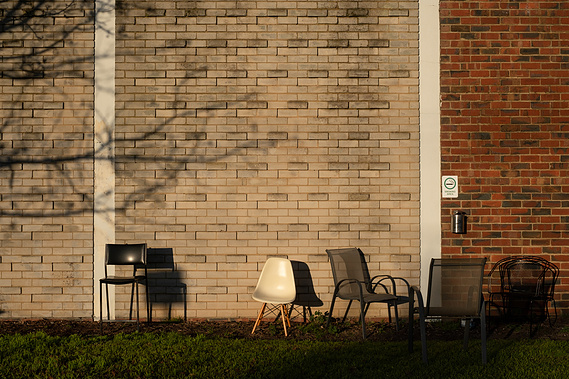 five chairs randomly placed on the grass in front of a red and blonde brick facade of an old factory with the shadow on of a tree with no leaves and a cigarette butt container and a sign saying 'smoking area'