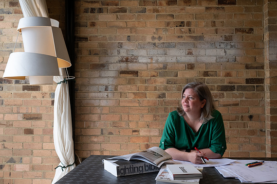 woman ( Adele Locke) in a long sleeved green blouse with her arms folded holding a pen. She's looking out to the left with books and papers in front of her on a steel desk. There is a handmade white lamp on the left and behind is a blonde brick wall