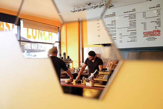 Man ( Marko Abramov) at the cash register handing a schnitzel roll to a customer in his Schnitzel takeaway food shop reflected in hexagon shaped mirrors on his wall. 