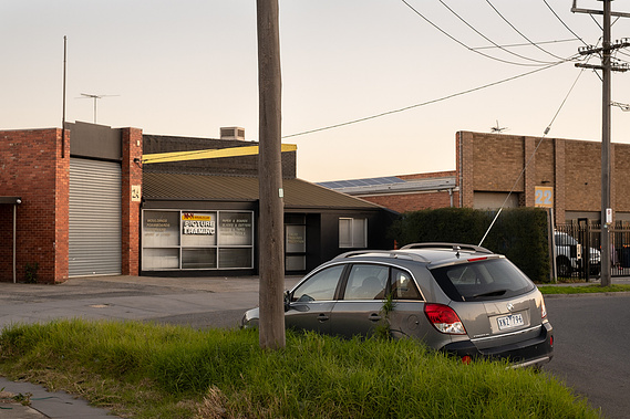 streetscape of factories with brick facades with power poles and long grass on the nature strip growing into the door of a parked door and snaking up the side