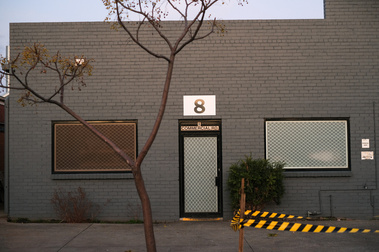 The facade of a 1950's brick factory painted a blue grey with a screen door in between 2 windows with aluminium screens and a tree in the front with some yellow and black tape Above the door it says '8 Commercial rd) and has a large no. 8 above that