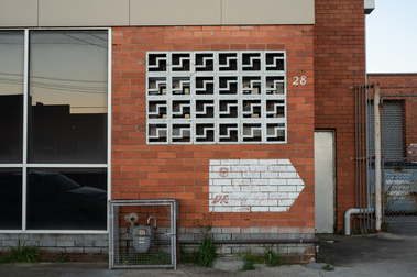 a 1950's red brick  factoryette facade with besser brick painted white  and other details including  the number 28,  a gas meter, weeds, fencing, a roller door, pipes and a reflection of power lines and another building at disk in the window