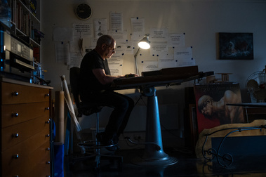 a man sitting  at an old drafting table, lit by a single lamp in an office surrounded by quotes and artwork