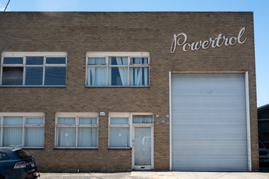 the facade of a 1950's light brown brick factoryette with peeling white paint on the window frames and a roller door and lettering saying 'Powertrol'. There is a station wagon in the front and blue sky above