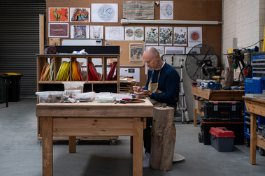 a man sitting at a large wooden workbench making mosaics in a neat factory with drawings pinned to the wall and tools and machinery along the wall