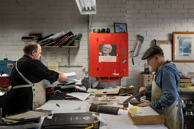 2 men on either side of a large work bench doing bookbinding, there are books and tools on the bench, a bookshelf with accounting folders and a red cupboard attached to the white wall that has a quote by warren buffet
