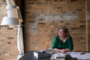woman in a long sleeved green blouse with her arms folded holding a pen. She's looking out to the left with books and papers in front of her on a steel desk. There is a handmade white lamp on the left and behind is a blonde brick wall
