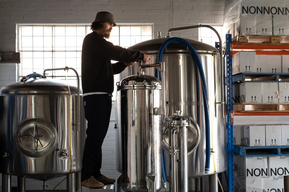 a man standing on a platform turning a wheel in a brewery with small and large fermentation vessels and a white brick wall with steel sash window in the background
