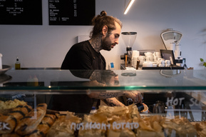 man with tattoos  in a deli pouring coffee, a coffee machine and menu in the background and a counter top with pastries in the front
