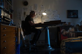 a man sitting  at an old drafting table, lit by a single lamp in an office surrounded by quotes and artwork
