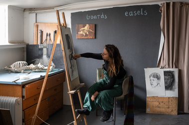 woman sitting painting at an easel in a studio that has a skeleton and paintings and the words 'easel' and 'boards'