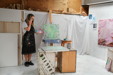 woman artist in her studio holding a paintbrush with white sheets pinned to the wall, painted white walls and floor, paintings on an easel and wall, she's working on a painting sitting on old tea boxes with the word 'fragile'