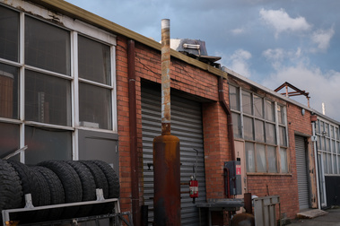 the side view of 1950's red brick factoryettes with a rack of old tyres, a chimney shaped object, roller doors and windows and blue sky with clouds