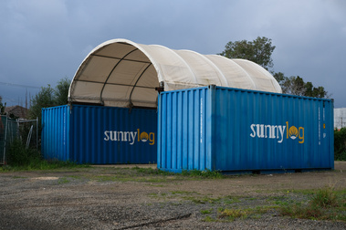 2 blue shipping containers on a cleared block with a dome shaped roof  structure over them. They have 'Sunny Log' written along the side