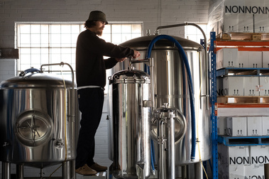a man standing on a platform turning a wheel in a brewery with small and large fermentation vessels and a white brick wall with steel sash window in the back ground
