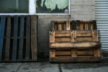 packing pallets leaning against a painted brick factory wall with windows , a roller door and peeling paint and a concrete slab on the ground