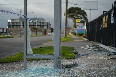 The frame of part of a bus stop with smashed glass, police tape and shattered glass on the ground with buildings and footpath