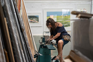 woman ( Catherine Jordan) picture framer using a machine with her foot on a pedal. There are pictures and painting frames leaning on the wall in the foreground and a white brick wall in the background with framed art work. 