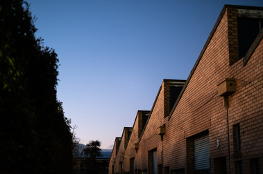 An old factory made of bricks with roller doors and sawtooth roof, blue sky at dusk and trees 